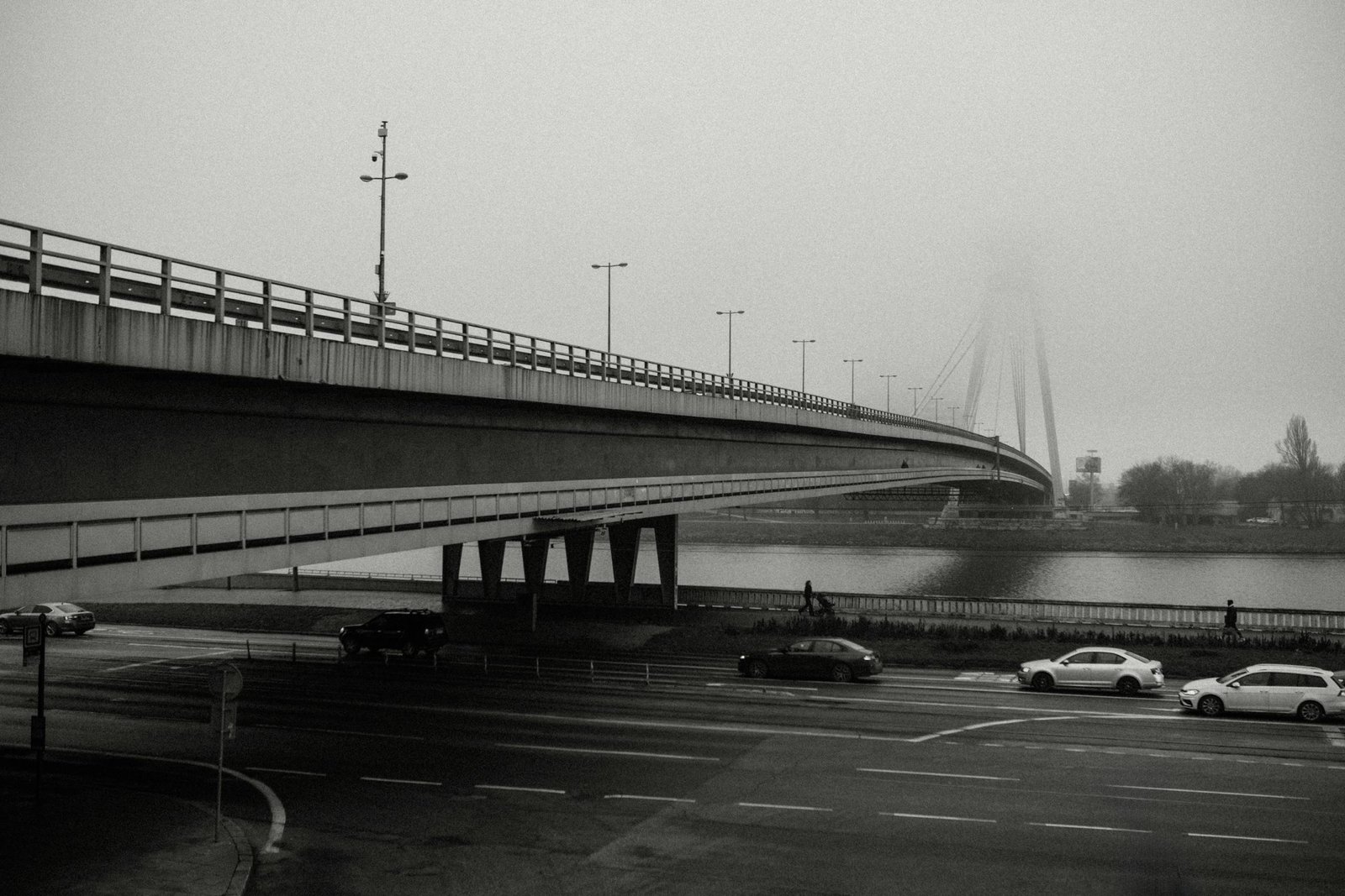 A misty morning view of a city bridge with light traffic and pedestrians, captured in black and white.