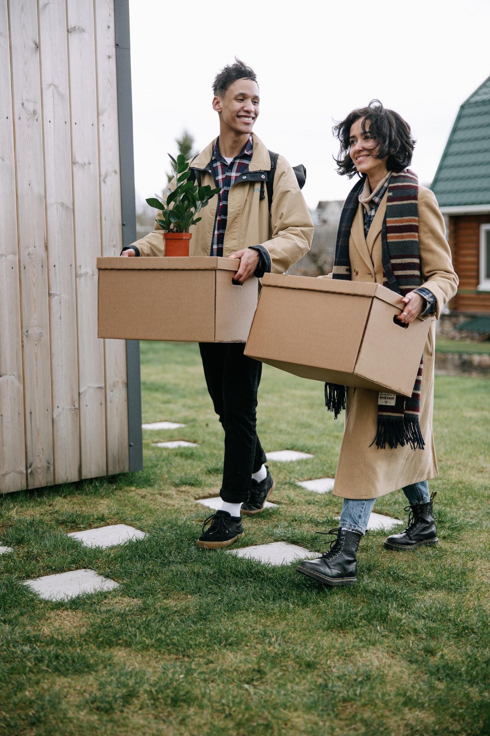 Happy couple carrying boxes and a plant while moving into their new home on a grassy lawn.